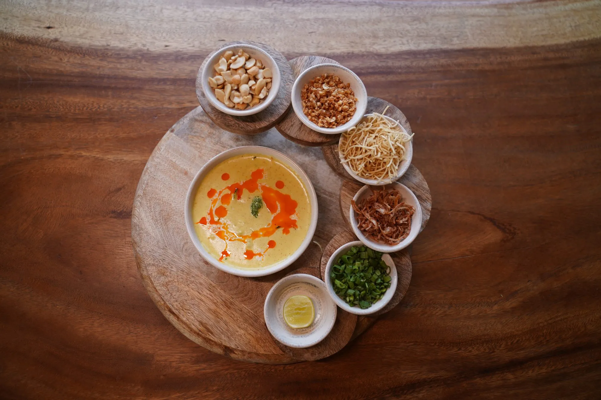 Small bowls of garnishes and a golden broth on a round wooden board