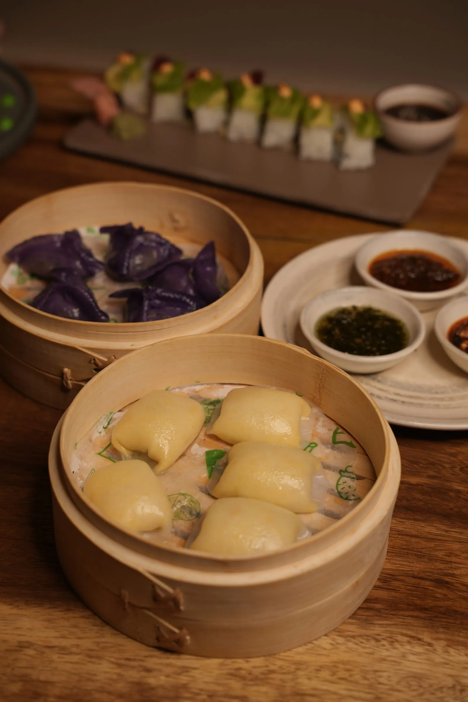 Bamboo steamers of purple and cream dumplings, with sushi rolls and dipping sauces behind