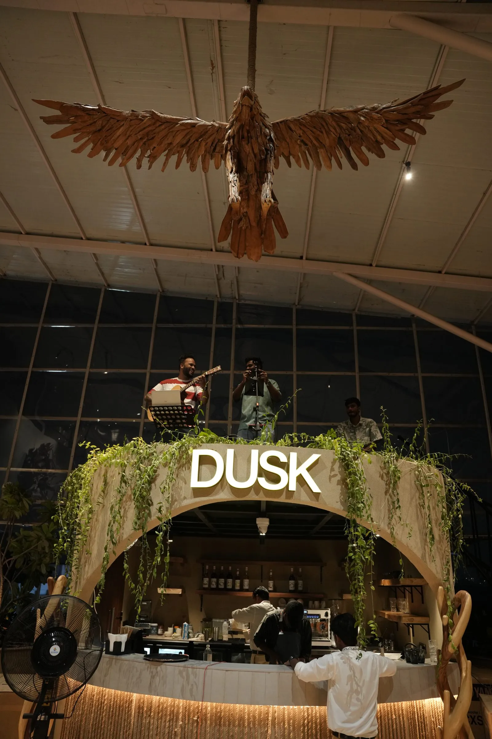 A band playing above the glowing DUSK arch at night, beneath the driftwood eagle